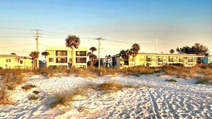 A row of yellow buildings with multiple stories, some with balconies, situated on a sandy area with sparse vegetation. - Bright, corner unit, with open floor plan and views from nearly every room!  (Bradenton Beach, Anna Maria Island)