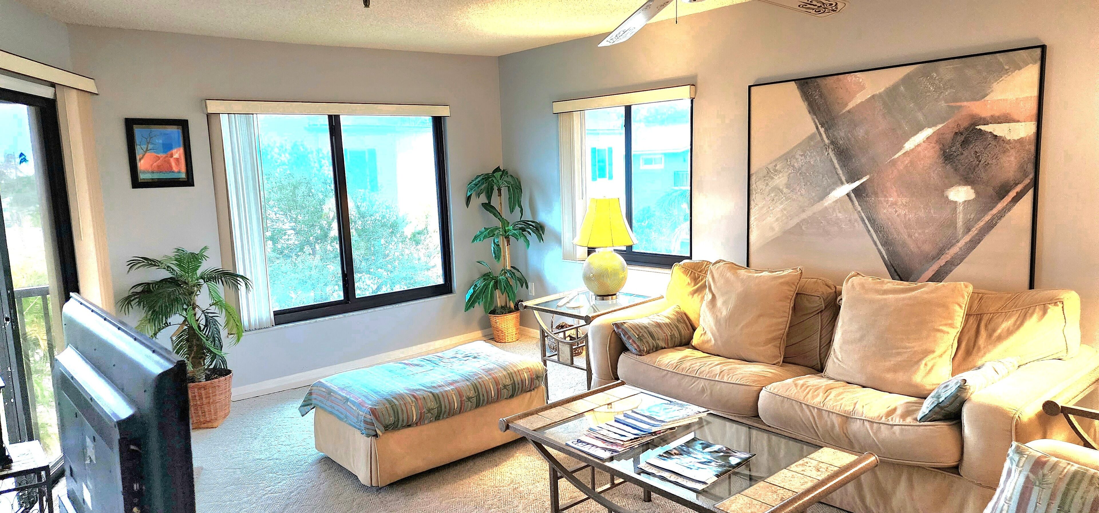 Una sala de estar moderna con sofá, reposapiés y mesa de centro, ventanas amplias y un cuadro en la pared.