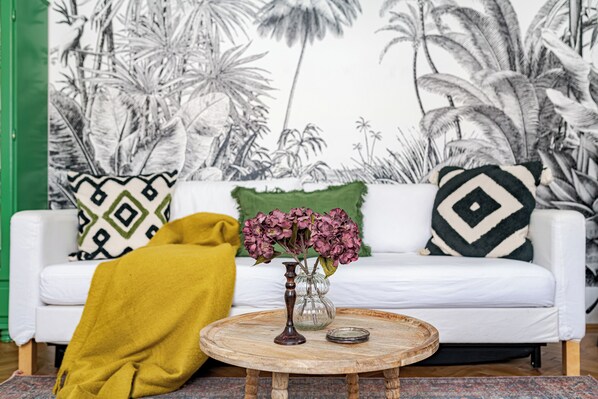 Un salón con sofá blanco, una mesa redonda de madera, un florero con flores y un mural tropical en la pared.