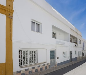 Um edifício branco com várias janelas e portas, uma varanda visível e uma placa com a inscrição 'QUARTEAS'.