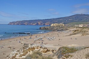 Una playa con gente tomando el sol y nadando, un litoral rocoso y edificios al fondo.