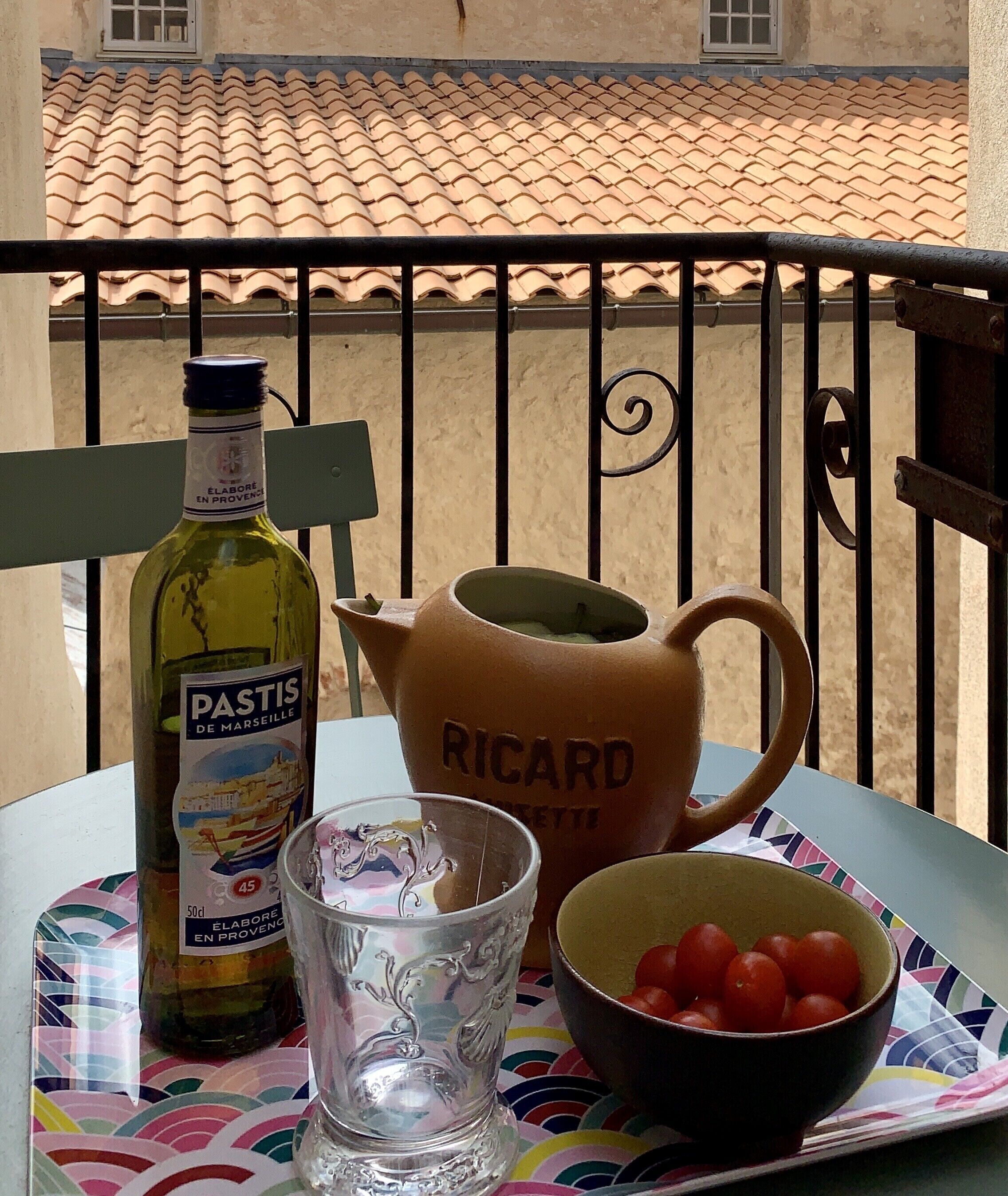 Et bord med en flaske Pastis de Marseille, et glas, en keramikkanne og en skål med kirsebærtomater på en balkon.