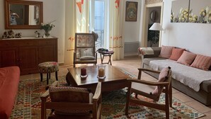 A cozy living room with a wooden coffee table, a sofa, a chair, and a dining table. There is a window with curtains, a mirror, and a wall-mounted artwork. - Large charming duplex, air-conditioned and WiFi in the heart of the citadel  (Bonifacio)