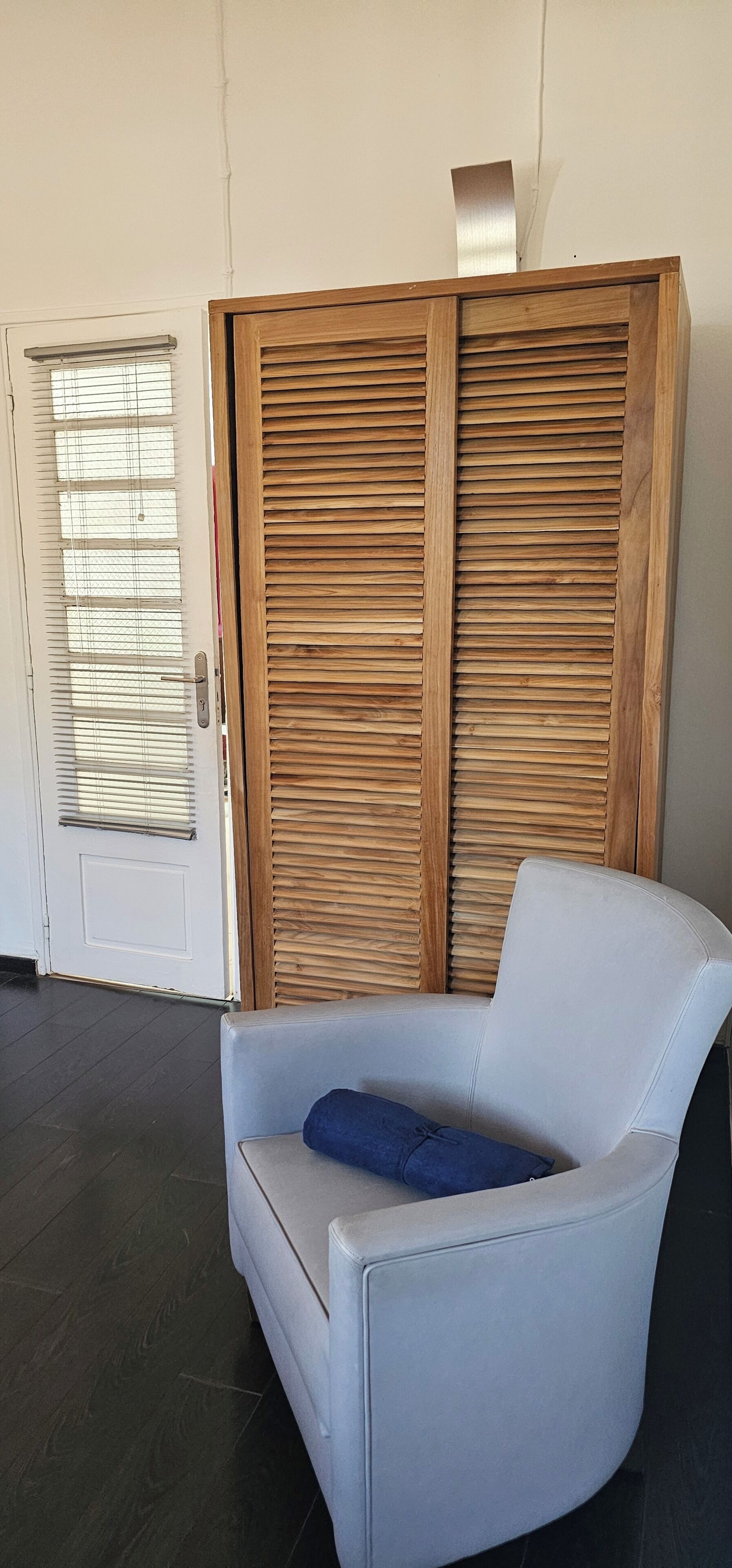 Ein Zimmer mit einer weißen Tür, einem Holzschrank und einem hellen Sessel mit blauem Kissen.