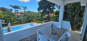 A balcony with a pool, outdoor seating, and a view of a lush garden and buildings.