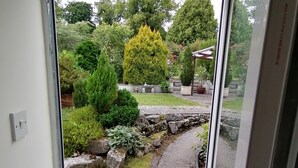 A view through a glass door into a garden with a stone pathway, a small water feature, and various plants. - Comfortable and spacious apartment in Middleton-by- Youlgrave near Bakewell.  (Middleton-by-Youlgrave)