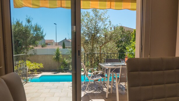 Udsigt fra et værelse med skydedør til en udendørs terrasse. Terrassen har en pool, et bord med stole og forskellige planter.