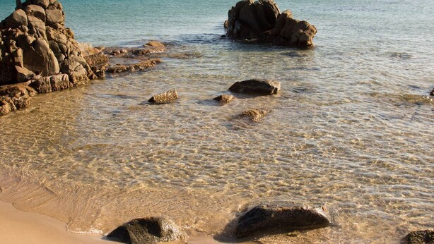 Ein Strand mit klarem Wasser, Felsen und einer entfernten Küste.