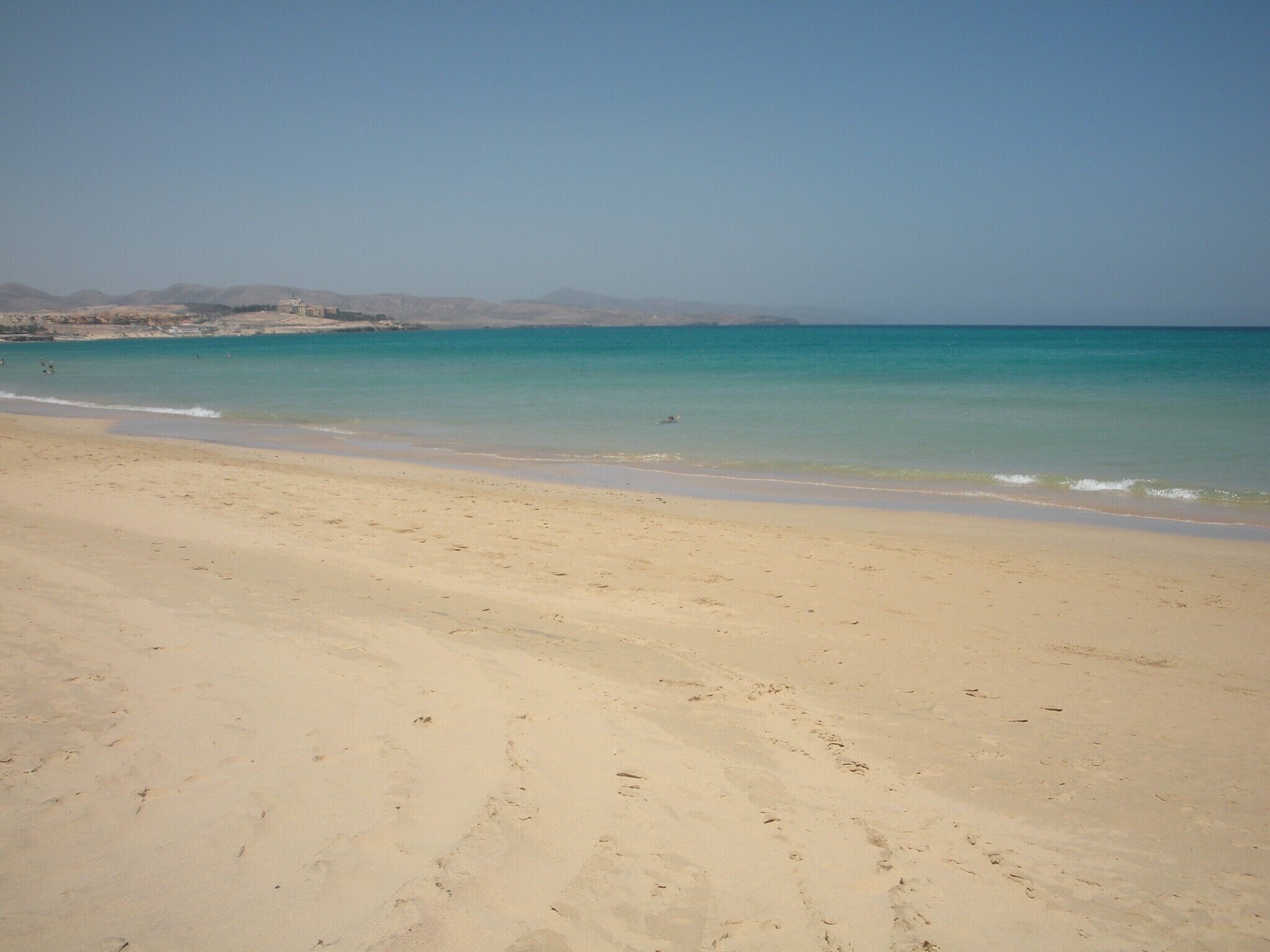 A wide sandy beach with gentle waves, a clear sky, and distant mountains.