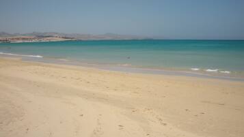 A wide sandy beach with gentle waves, a clear sky, and distant mountains.
