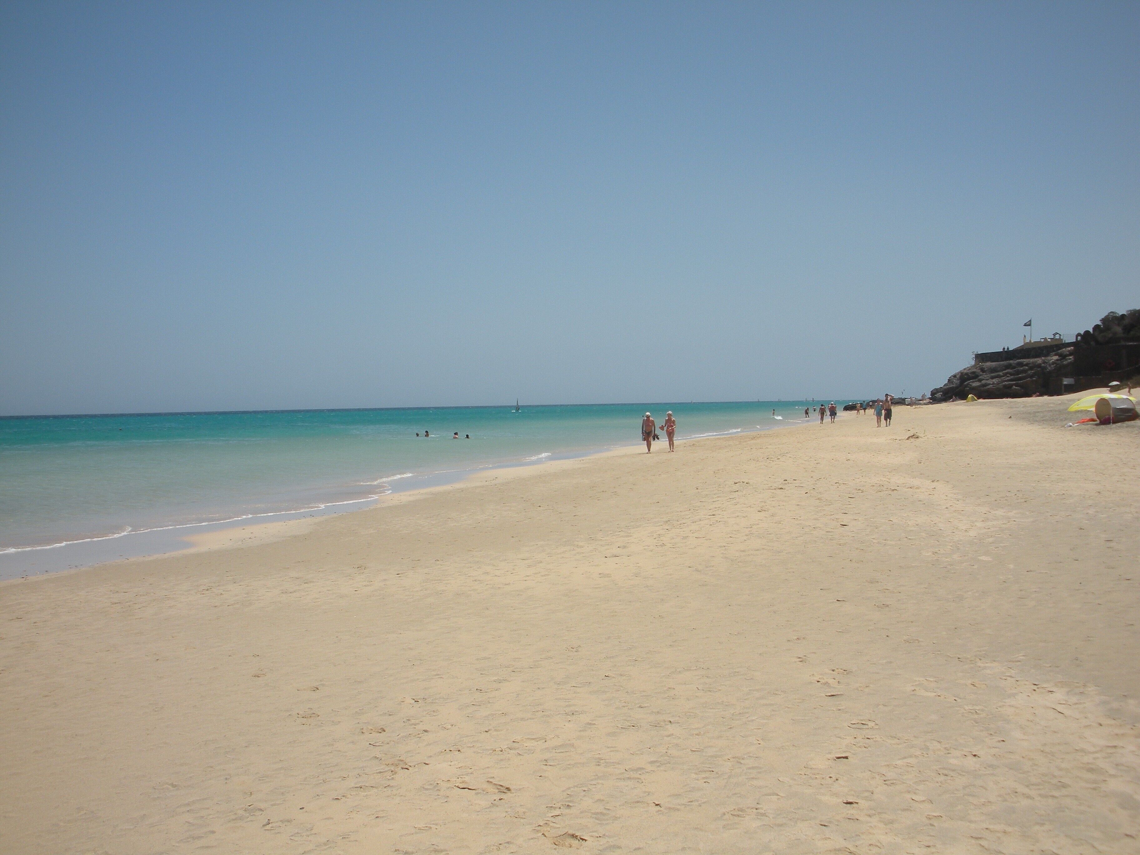 Een breed zandstrand met helder turquoise water, enkele mensen die wandelen, en een rotsachtige klif aan de rechterkant.