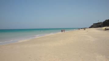 Een breed zandstrand met helder turquoise water, enkele mensen die wandelen, en een rotsachtige klif aan de rechterkant.
