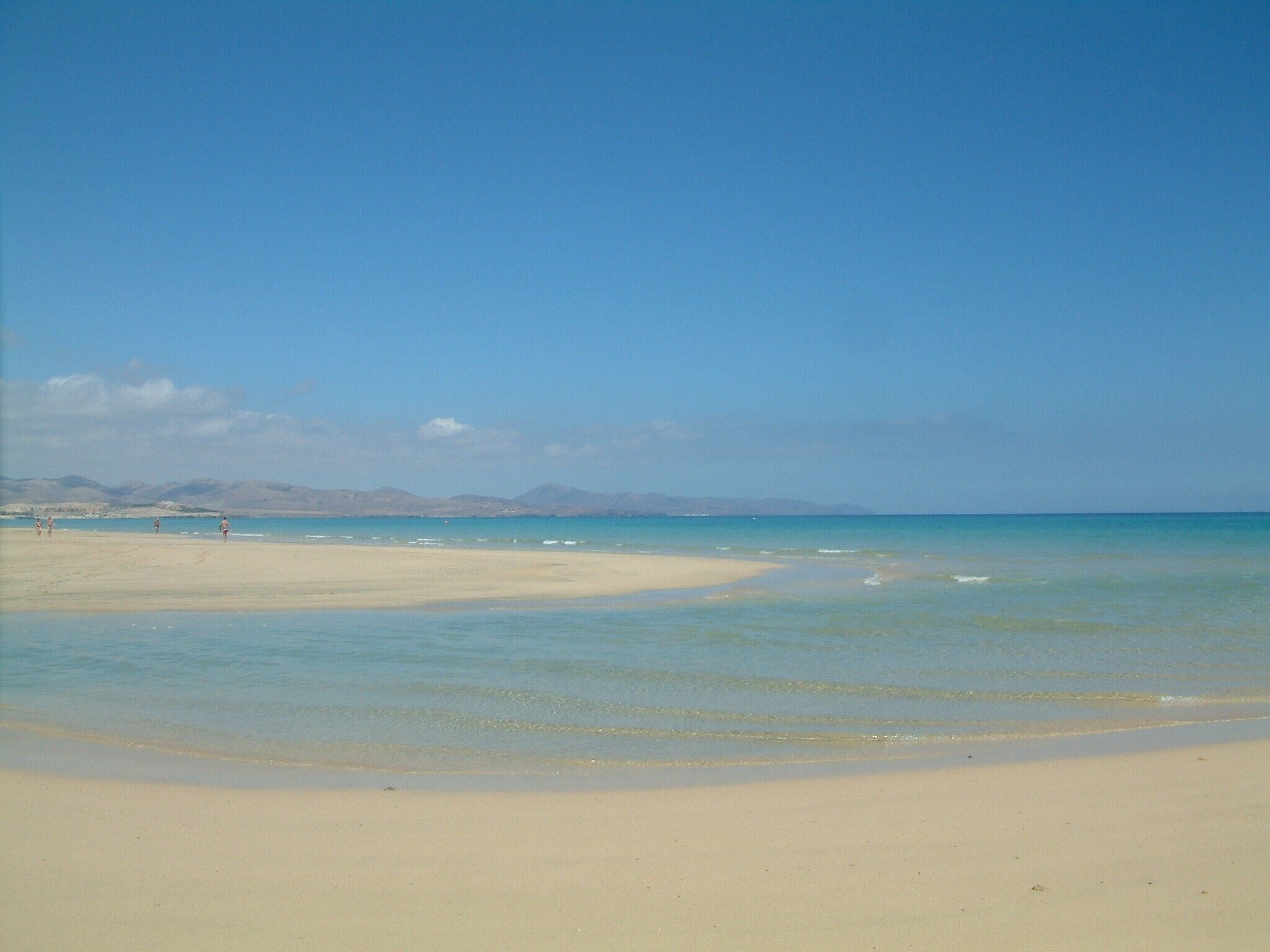 Een strand met helder water, een zandstrand en bergen in de verte.