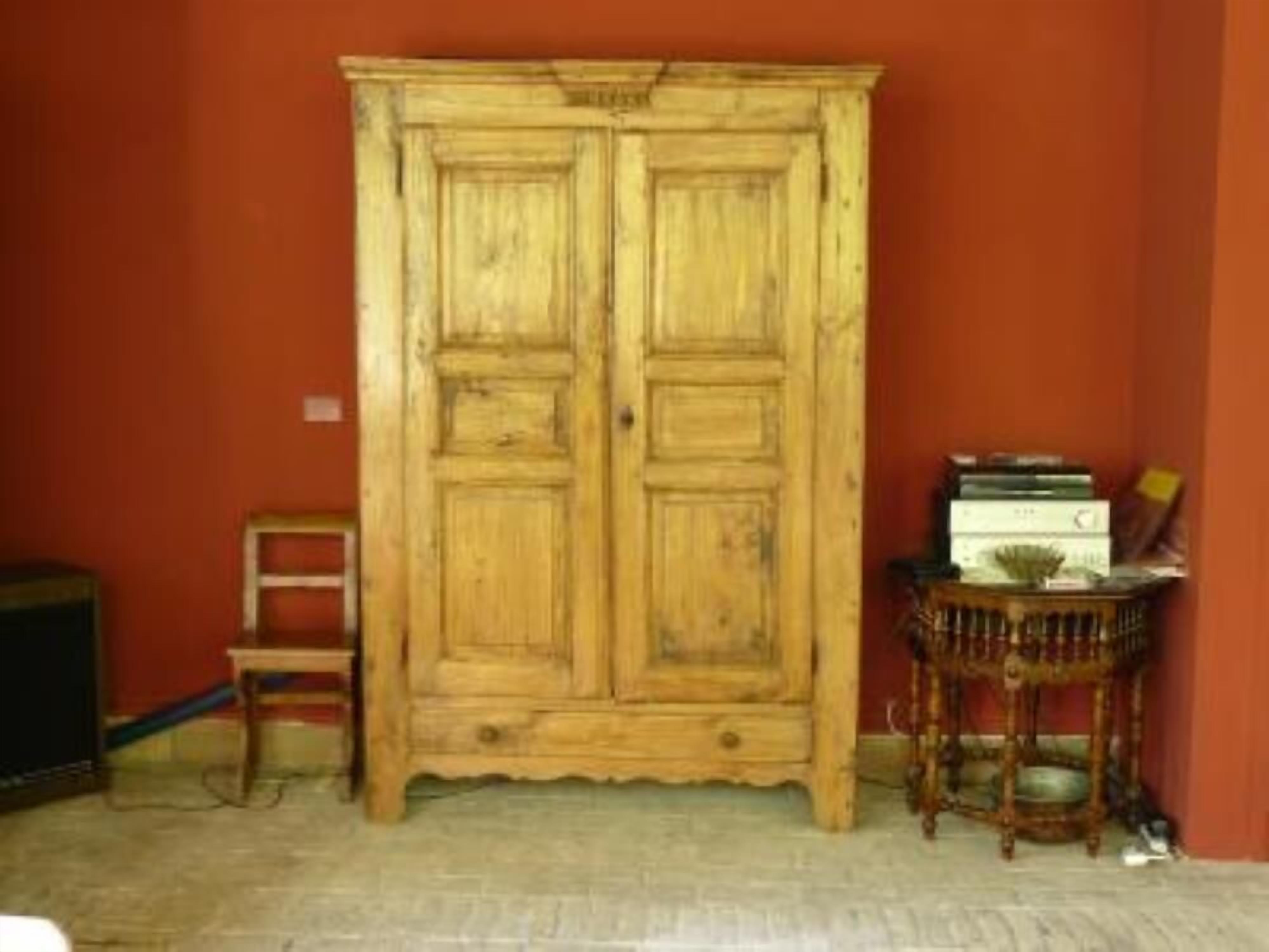A wooden wardrobe with two doors, a small table with books, and a chair in a room with red walls.