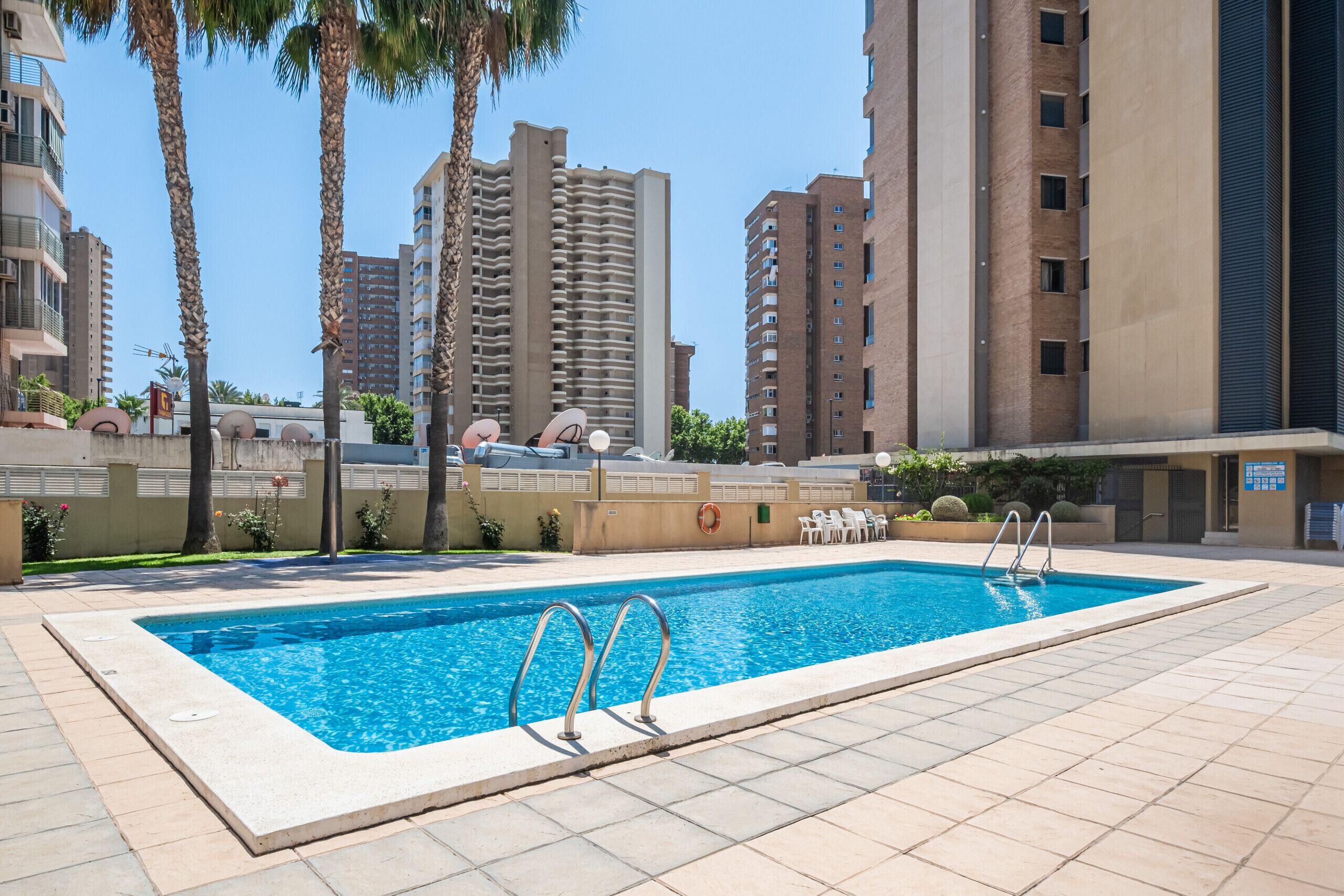 Uma piscina retangular com escadas de metal, rodeada por um deck ladrilhado. Edifícios altos e palmeiras aparecem ao fundo.