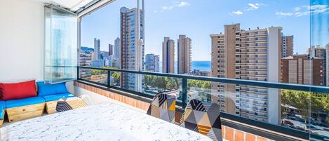 A balcony with a city view, a bed with patterned bedding, and a seating area with cushions and geometric-patterned chairs.
