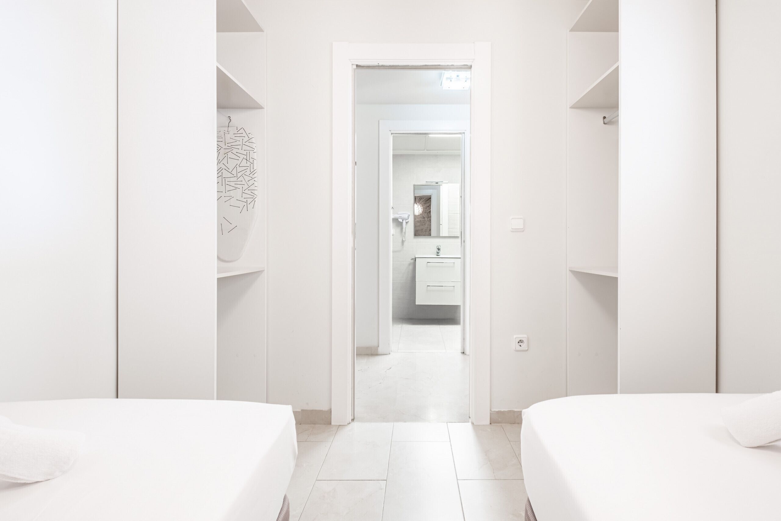 A hotel room with a clean, minimalist design, featuring two beds, a bathroom with a mirror, and a white color scheme.