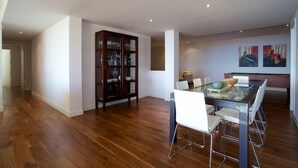 Un comedor amplio con una mesa de vidrio y sillas blancas, un mueble de madera con puertas de cristal, y dos cuadros en la pared.