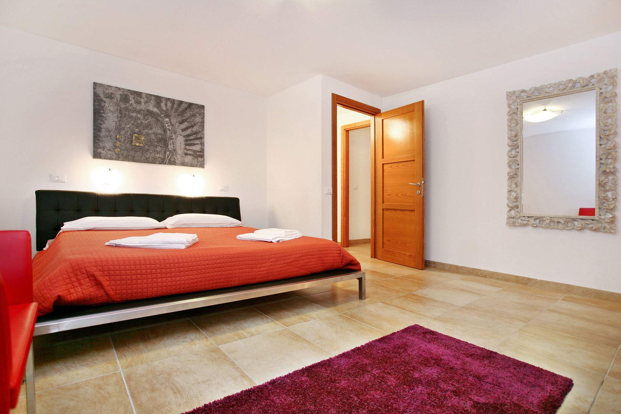 Un dormitorio con cama, una silla roja, una puerta de madera y un espejo en la pared.