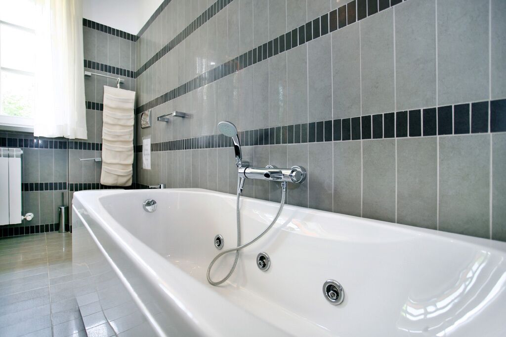 A modern bathroom with a large bathtub, grey and black tiled walls, and a white towel hanging on a rack.