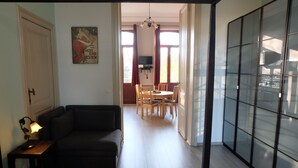 A room with a bed, a dark sofa, a small table with a lamp, a dining area with chairs and a table, and a large sliding glass door with a view.