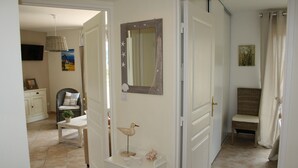 Un couloir avec une porte blanche, une chaise en osier, une petite table ornée d’objets décoratifs et un miroir accroché au mur.