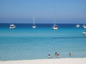 Ein Strand mit klarem türkisblauem Wasser, mehreren Segelbooten und einigen badenden Personen.