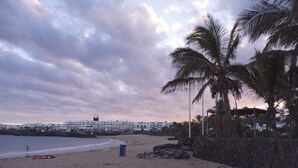 A beach with a stone wall, palm trees, and a row of white buildings in the background. - Thomas Beach House Costa Teguise (Costa Teguise)