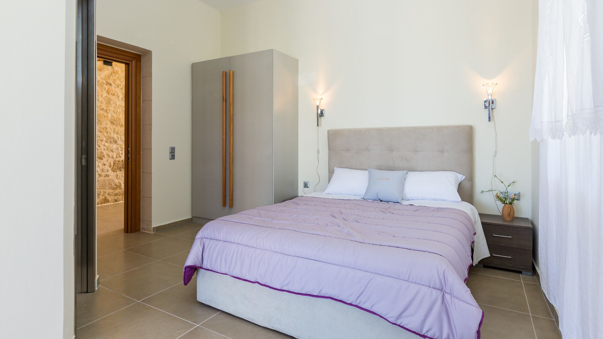 A bedroom with a bed, bedside tables, a nightstand, a wall-mounted lamp, and a door leading to another room.