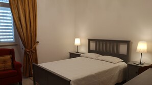 Une chambre à coucher bien rangée, avec un lit, deux tables de chevet dotées de lampes, un fauteuil rouge et une fenêtre avec des stores.