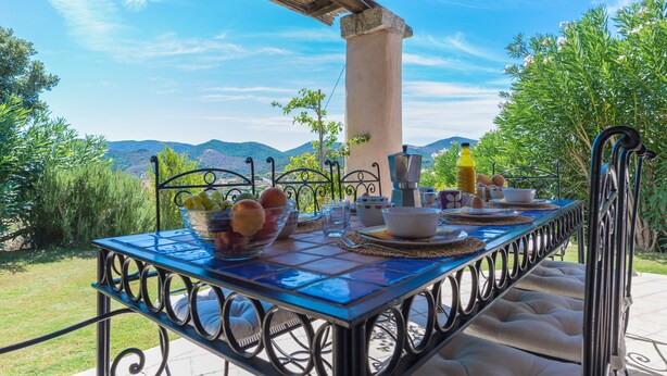 An outdoor dining table with a view of the mountains, set with breakfast items and a bottle of orange juice.