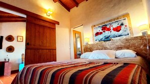 Ein Schlafzimmer mit einem Holz-Kopfende, einem Bett mit weißer Bettwäsche, einer farbenfrohen, gestreiften Tagesdecke, einem Bild von roten Blumen an der Wand und einer Holztür.