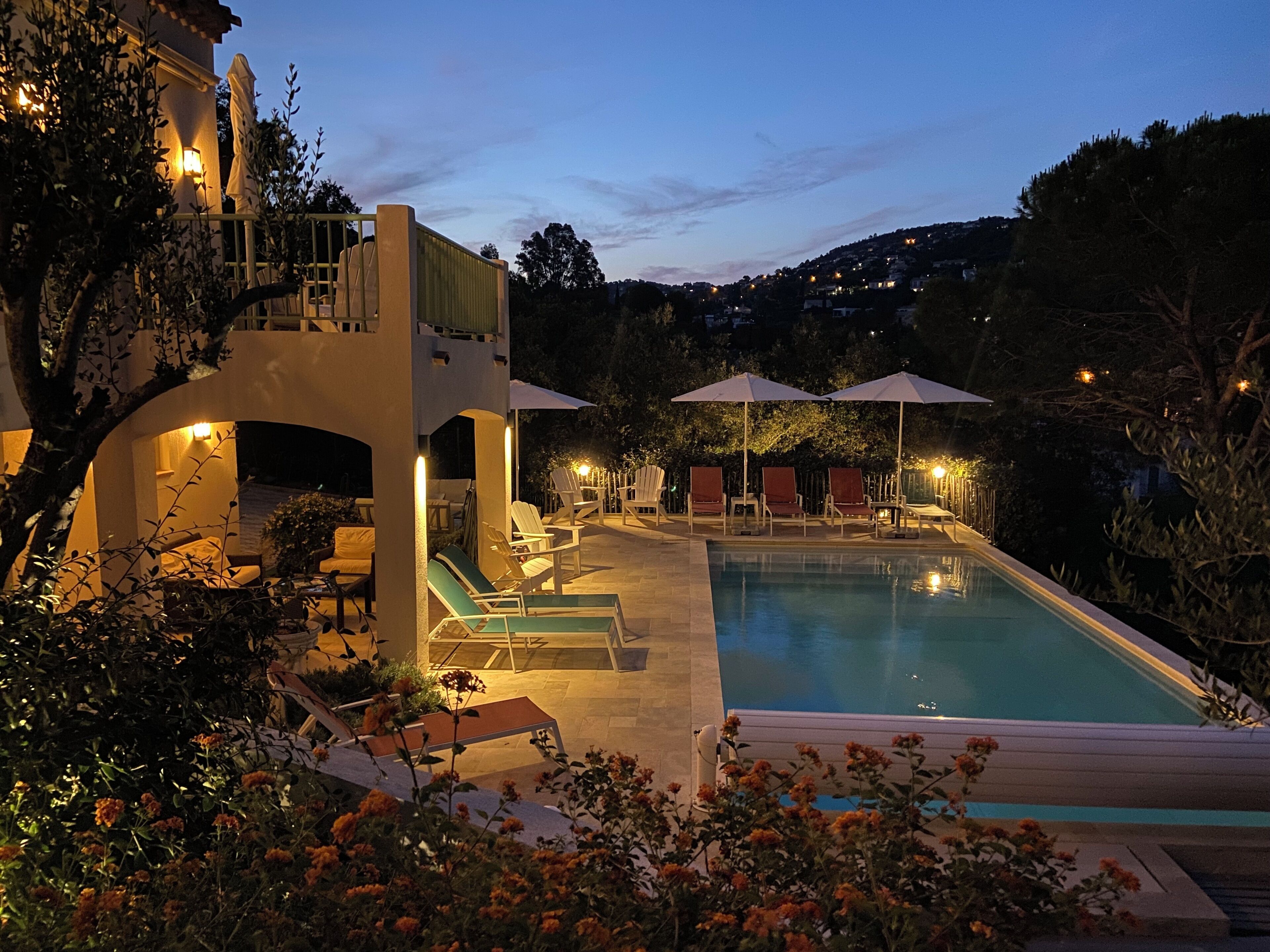 A pool area with lounge chairs and umbrellas, surrounded by greenery and a building with a balcony.