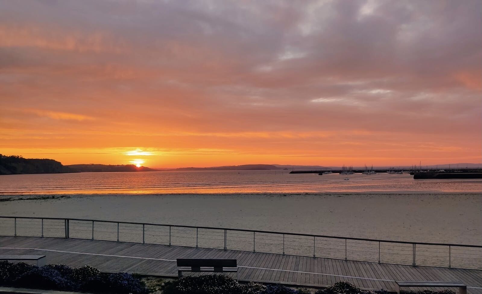 Un coucher de soleil sur une plage, avec une promenade en bois et un banc.