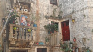 Un carrer estret amb un edifici de pedra que té un balcó amb plantes en testos, una porta vermella i una finestra petita.