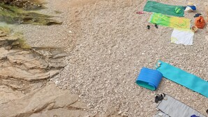 Una platja de petits pedres amb aigua cristal·lina, un promontori rocos i una illa a la distància.