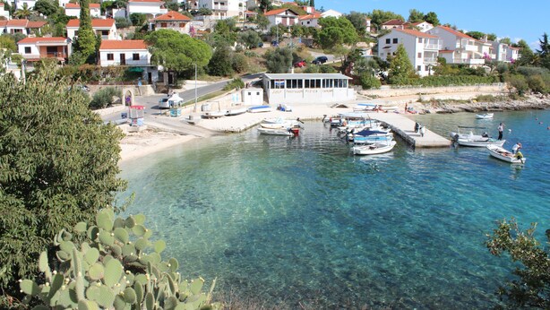 Un poble costaner amb aigües cristal·línes, vaixells amarrats en una marina i edificis residencials amb teulades vermelles.