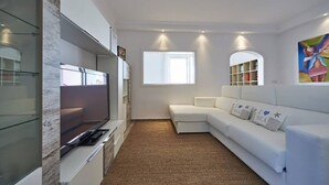 Ein modernes Wohnzimmer mit Flachbildfernseher, einem weißen Sofa, einem Bücherregal und einem großen Fenster.