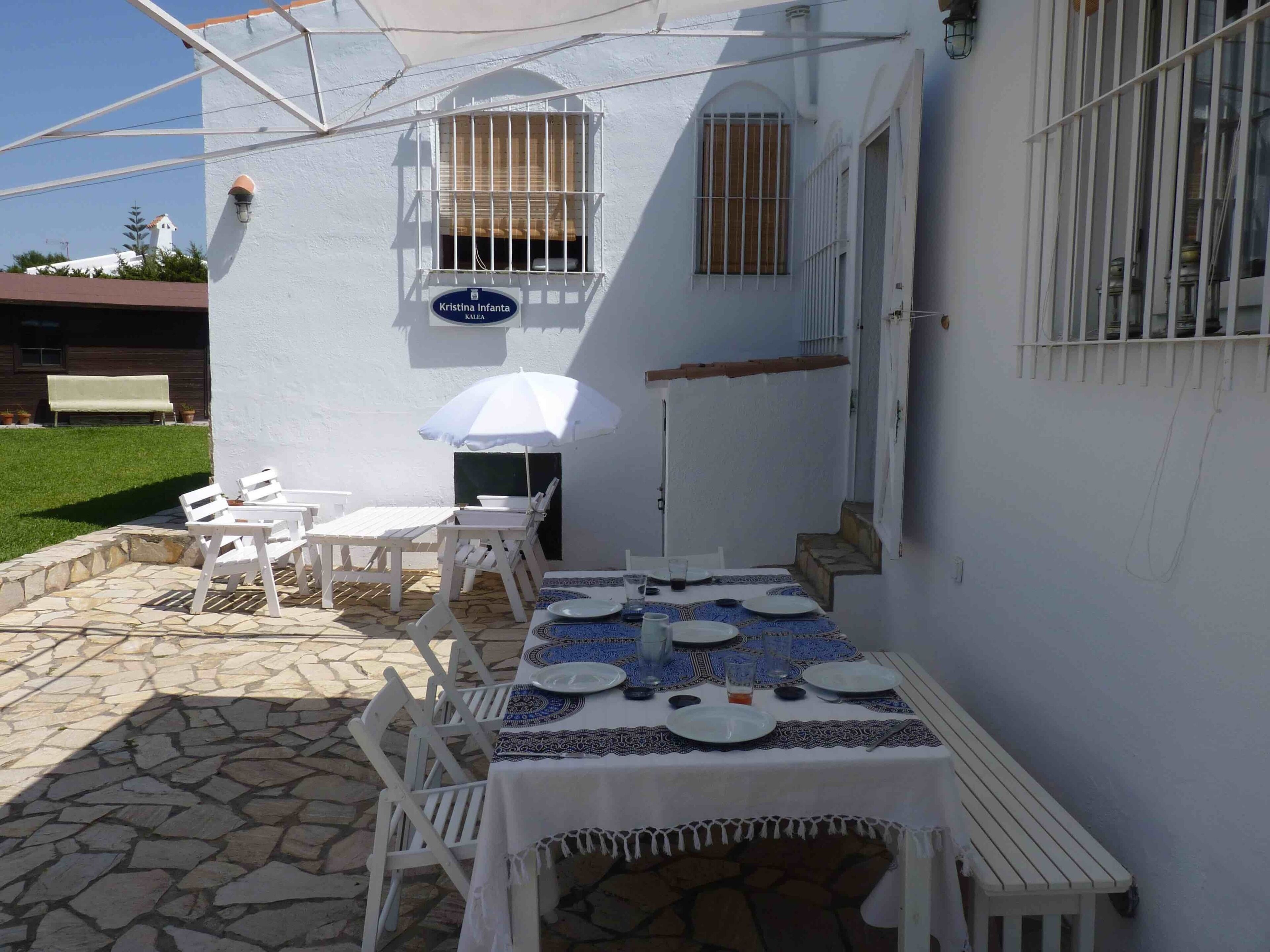 An outdoor patio with a table set for dining, white chairs, and a white umbrella.