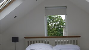 Ein Schlafzimmer mit schrägen Wänden, einem Fenster mit Blick auf Bäume, einem Bett mit weißen Leinen und zwei gefalteten Handtüchern, einem Nachttisch mit Lampe und einem Beistelltisch mit Telefon.
