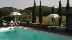 Piscina con sillas de descanso y sombrillas, rodeada de árboles y colinas.
