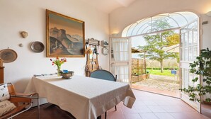 Un comedor con una mesa, sillas, un cuadro y vistas al jardín a través de un ventanal abierto.