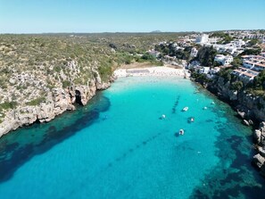 An aerial view of a coastal town with a clear turquoise bay, rocky cliffs, and white buildings with red roofs. - Apartment with dream view in Cala en Porter (Self check-in/out) (Cala 'n Porter)
