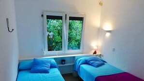Una habitación con dos camas, una ventana que da a zonas verdes, una lámpara en la pared y una mesita con lámpara.