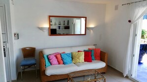Una sala de estar con un sofá blanco, cojines coloridos, una silla de mimbre, una mesa de centro de vidrio y un espejo en la pared.