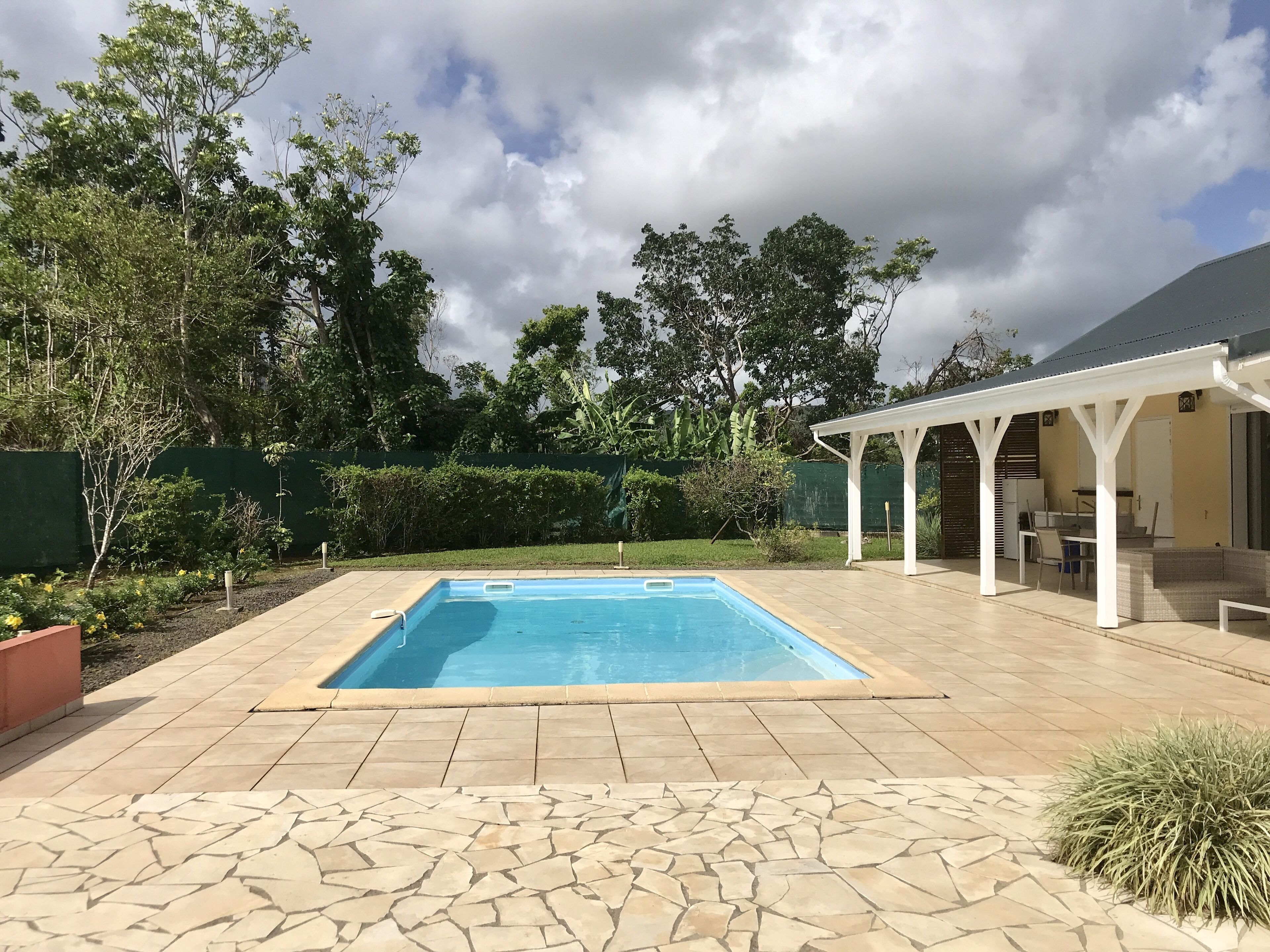Une piscine rectangulaire dotée d’une terrasse carrelée, entourée de verdure et agrémentée d’un patio couvert.