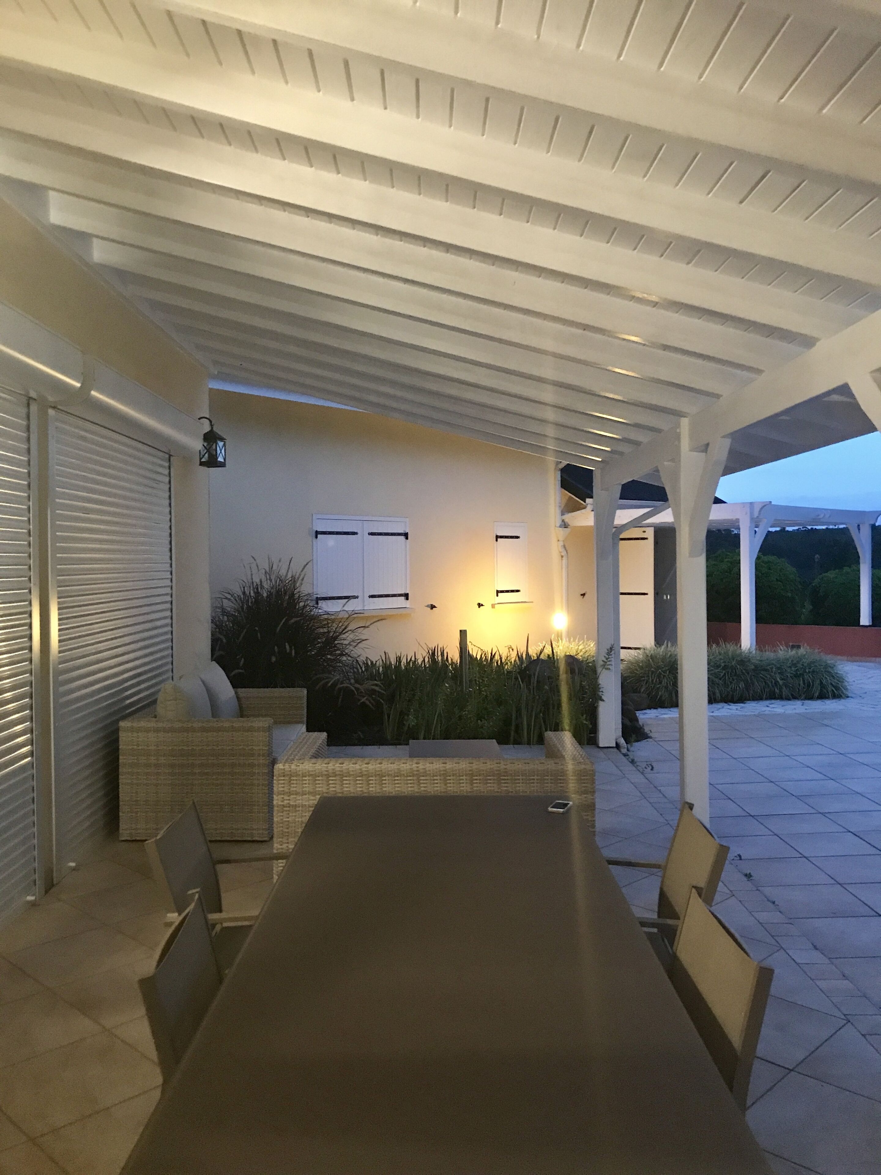 A covered outdoor dining area with a long table and chairs, a pergola overhead, and a wall with a window and plants.