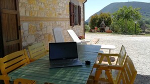 Ein Steinhaus mit einem Laptop auf dem Tisch, Außenbereich zum Sitzen und Blick auf einen Hügel.
