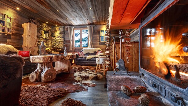 Interior rústico de una cabaña con pared de piedra, techo de madera y chimenea con leña ardiendo.