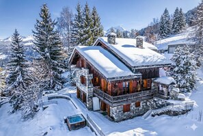Een besneeuwde lodge met een gevel van natuursteen, houten balkons en een jacuzzi op de voorgrond.